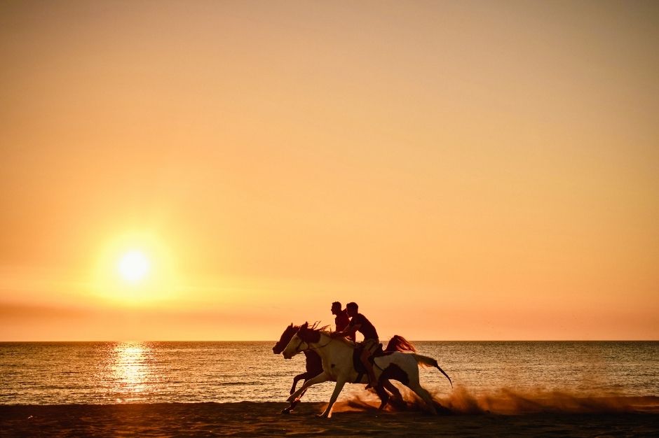 Two people on horseback galloping along the beach at sunset